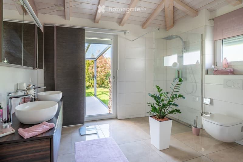 Modern bathroom with double sinks, a large mirror, a glass-enclosed shower, and a potted plant. The space features light-colored tiles and wooden accents.