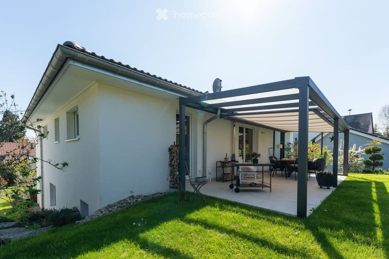 A modern patio area attached to a house, featuring a covered pergola, outdoor furniture including a table and chairs, and a well-maintained lawn.
