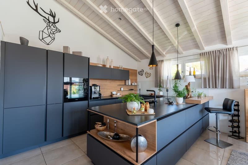 Modern kitchen with grey cabinets, wooden accents, and a central island. Features include a coffee machine, pendant lights, a wine rack, and decorative items on the island and shelves.