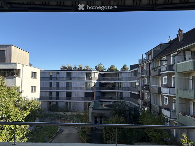 View from a balcony overlooking a multi-story residential complex with numerous balconies, trees, and a paved pathway.