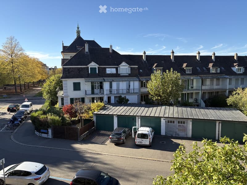 Multi-story residential building with a garage and parking area, surrounded by trees and parked cars.