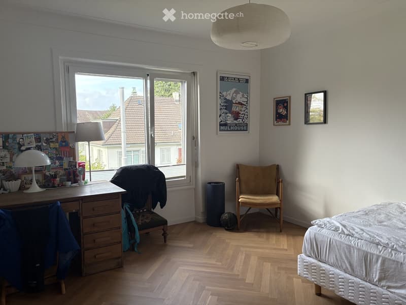 Bright bedroom with wooden flooring, large window, bed, desk, chair, and various decor items.