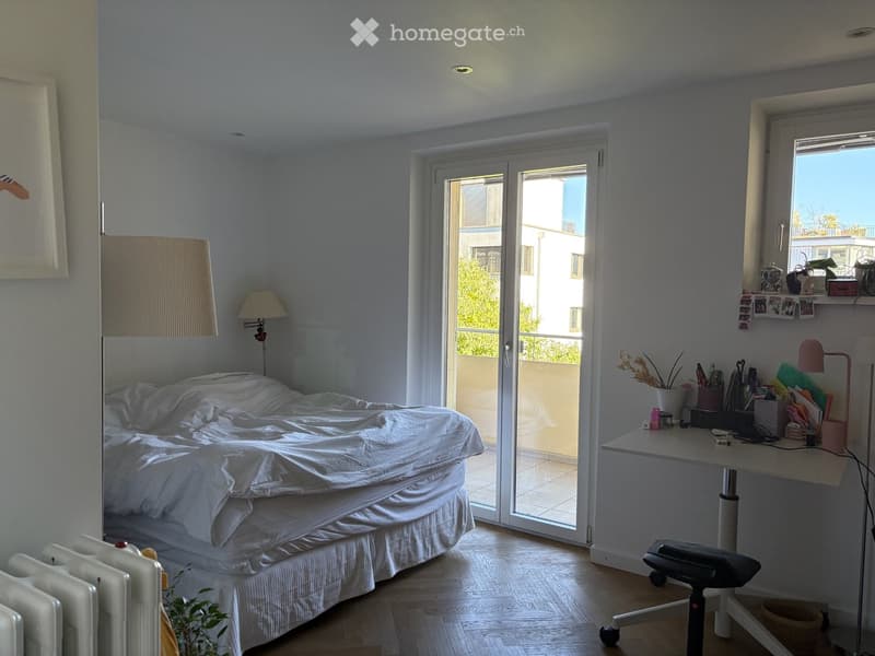 Bright bedroom with a bed, desk, chair, and radiator. Large window and sliding glass door lead to a balcony.