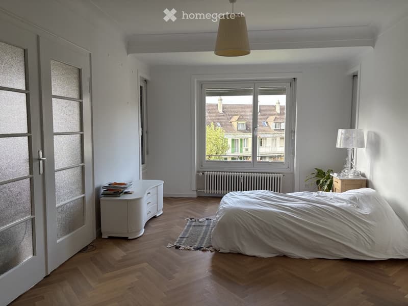 Bright bedroom with wooden flooring, large window overlooking rooftops, bed with white bedding, nightstand with lamp, small table with books, and a plant near the window.