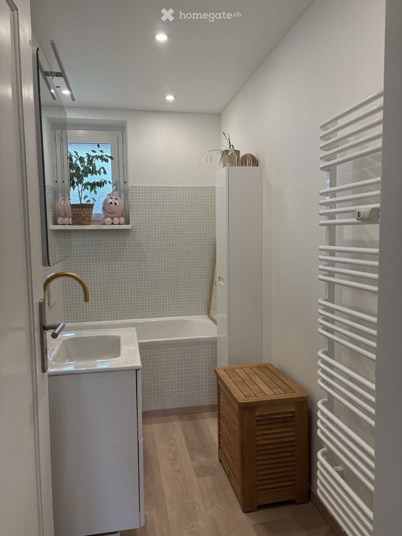 Modern bathroom with a white sink, gold faucet, bathtub, wooden stool, radiator, and decorative items on the windowsill.
