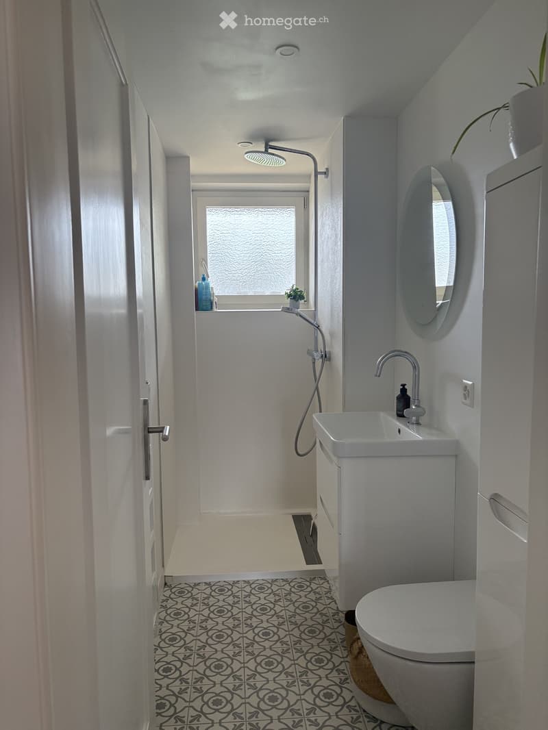 Modern bathroom with white walls, black and white patterned tile floor, toilet, sink, mirror, and shower area with a glass window.