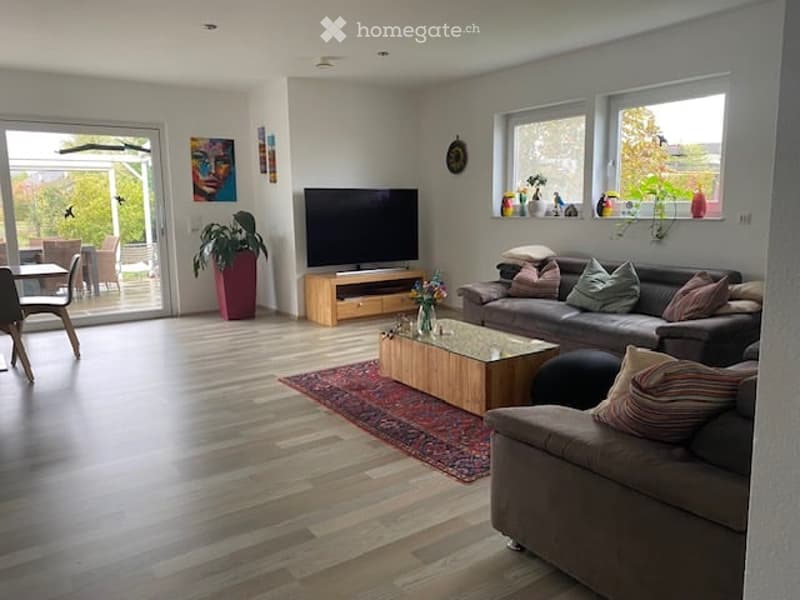 Bright living room with wooden flooring, large windows, a grey sofa, coffee table, TV, and dining area visible through the open sliding doors.