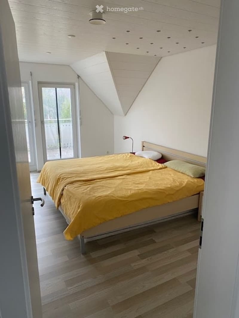 Bright bedroom with wooden flooring, large window with balcony access, neatly made bed with yellow bedding and pillows, and a bedside lamp.