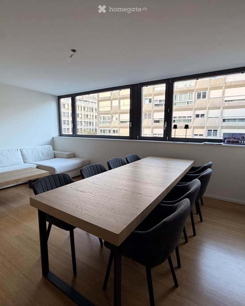 Modern, minimalist room with a white sofa, coffee table, large wooden dining table, and several chairs. Large windows offer a view of the city.