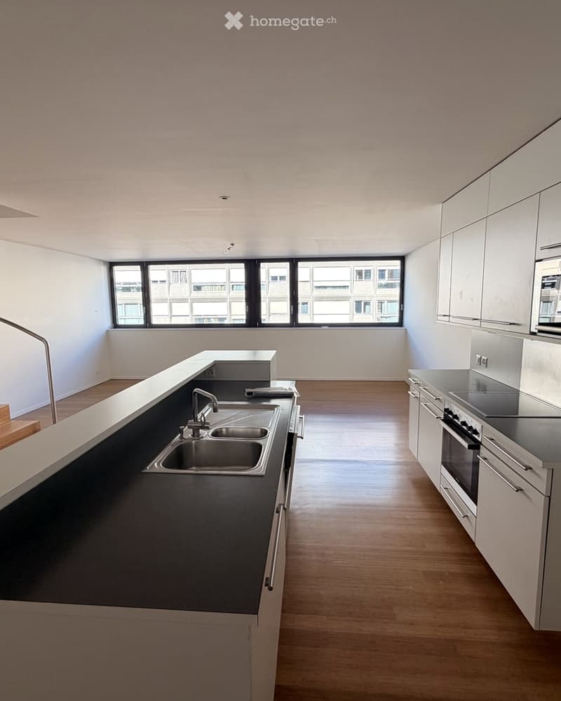 Empty kitchen with white cabinets, dark countertops, a sink, and a stove. The room features wooden flooring and large windows offering a view of the outside.