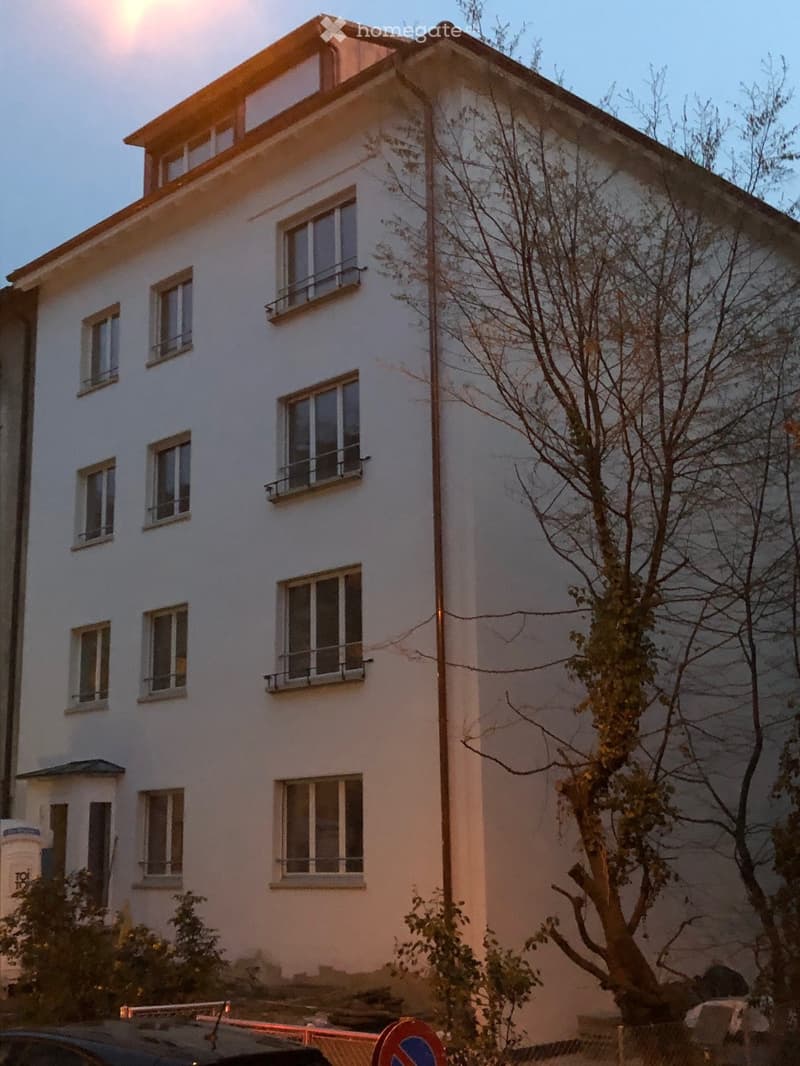 A two-story white house with multiple windows and balconies, surrounded by trees and a parked car.