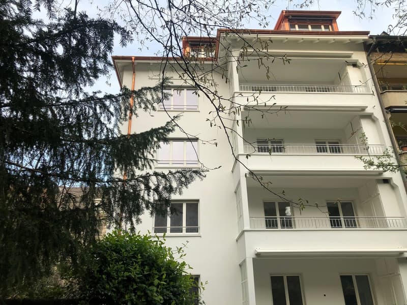 A multi-story white building with balconies and large windows, partially obscured by trees and foliage.