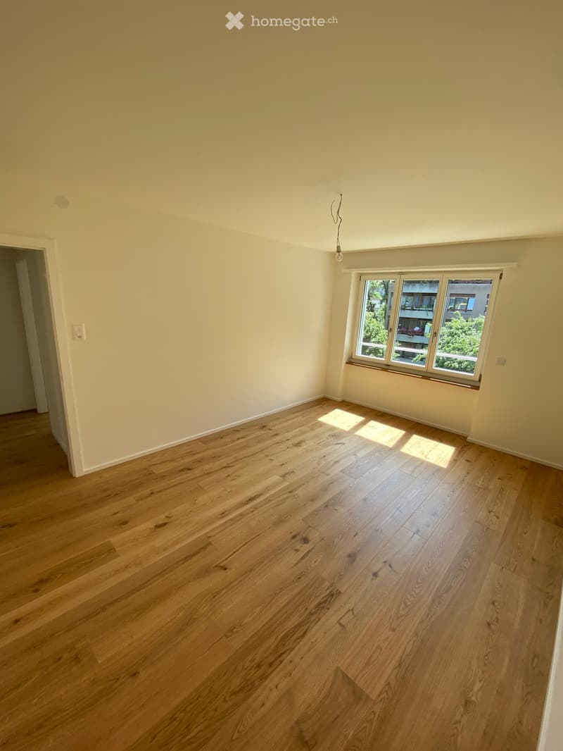 Empty room with light wooden flooring and white walls, featuring a large window with a view of greenery and a neighboring building.