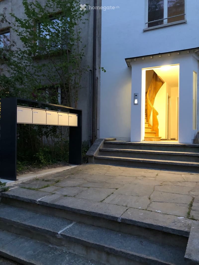 Modern two-story house with light blue exterior, illuminated entrance, stone steps, and a small garden area with a mailbox.