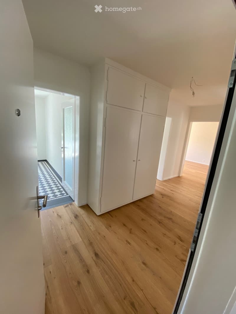 Empty hallway with light wooden flooring, white walls, and a built-in white cabinet. The hallway leads to other rooms with visible checkered tile flooring in one direction.
