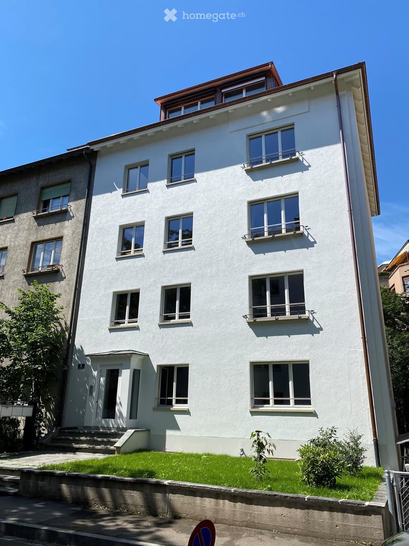 A modern, three-story residential building with white walls, multiple windows, and balconies. The building has a flat roof with a small attic section and is surrounded by a small garden area with green grass and shrubs.