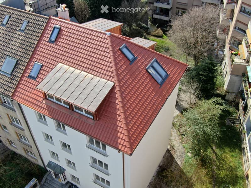 A multi-story residential building with a red-tiled roof, multiple windows, and a small flat roof section with skylights.