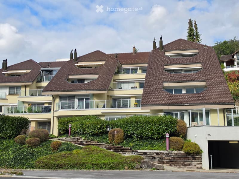 A large, multi-story house with a brown tiled roof, multiple balconies, and a well-maintained garden with shrubs and trees.