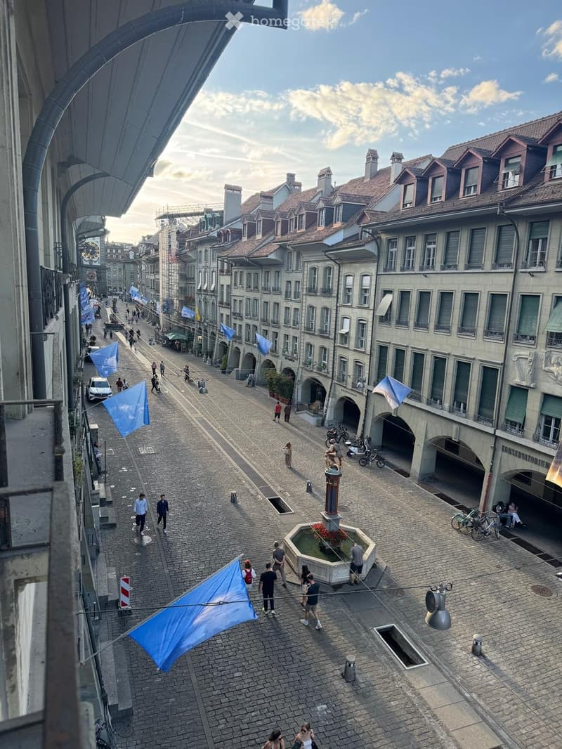 A cobblestone street lined with historic buildings, featuring a central fountain, pedestrians, bicycles, and flags hanging from wires.