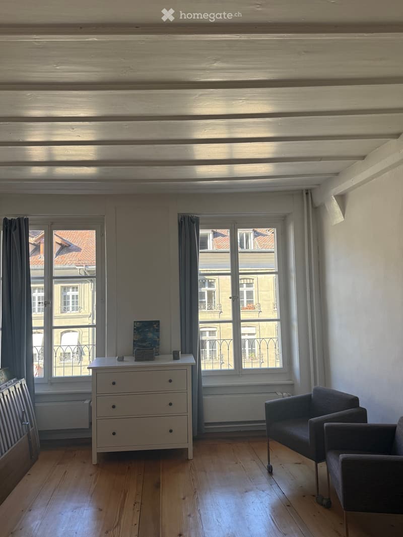 Bright room with wooden flooring, large windows, a small table with a cup and picture frame, and two chairs.