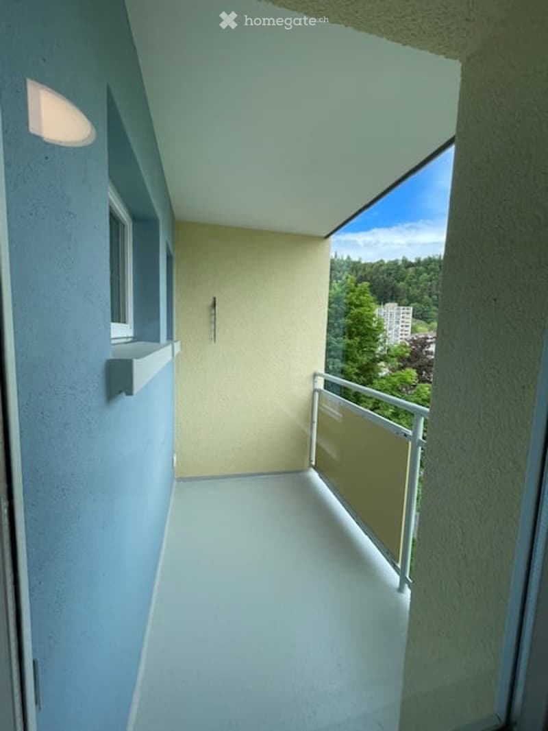 Balcony with white flooring, glass railing, and a view of trees and a building.