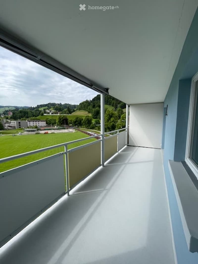 Spacious balcony with white flooring, metal railings, and large windows offering a scenic view of green fields and distant hills.