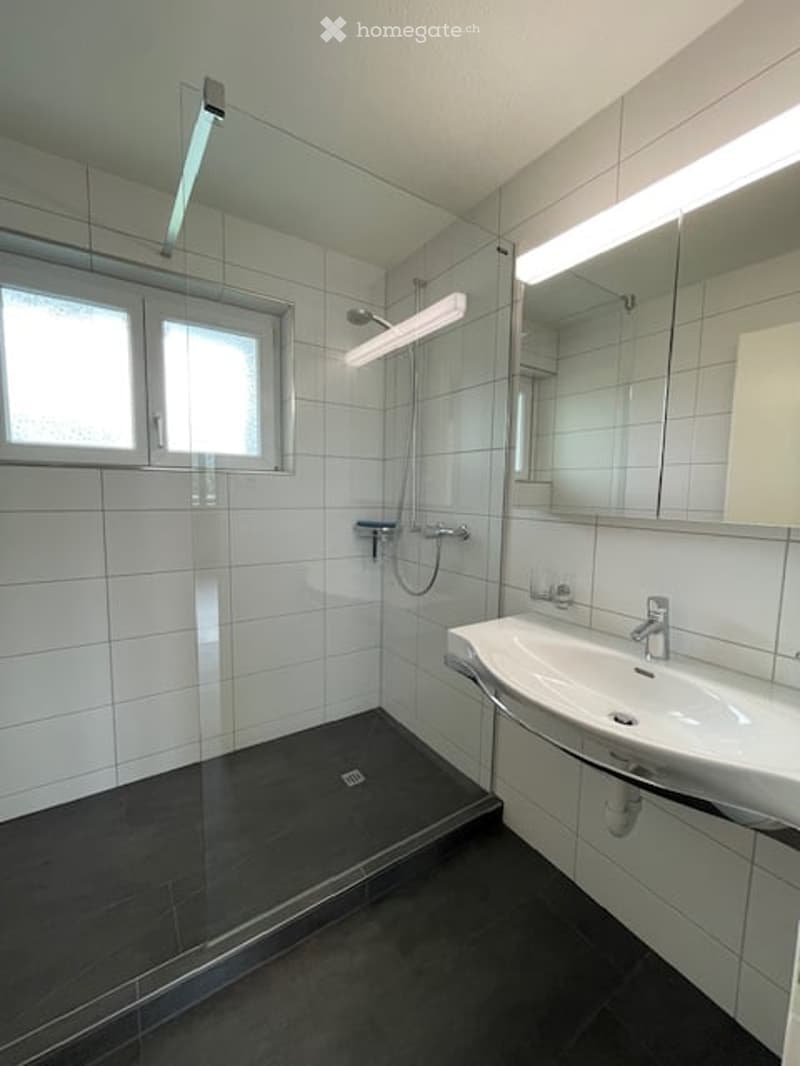 Modern bathroom with white tiles, a glass-enclosed shower, a white sink with a mirror, and a window for natural light.