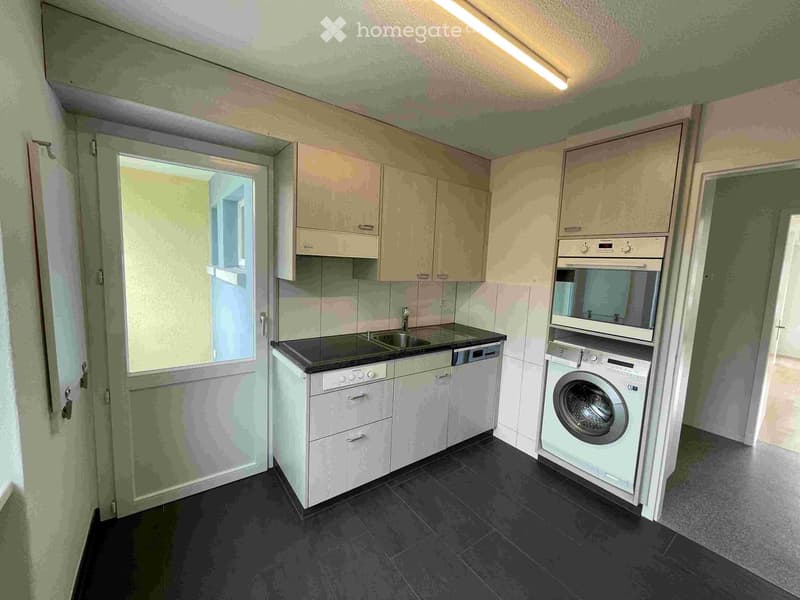 Modern kitchen with white cabinets, black countertop, sink, dishwasher, washing machine, and oven. The space is well-lit with a window providing natural light.