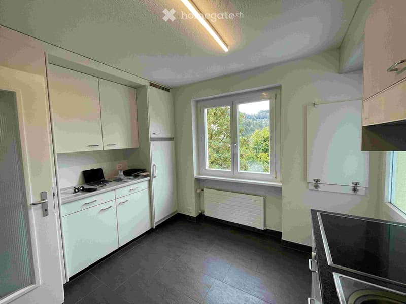A modern kitchen with light green cabinets, a black tiled floor, and a window overlooking greenery. A laptop is placed on the counter along with some kitchen items.