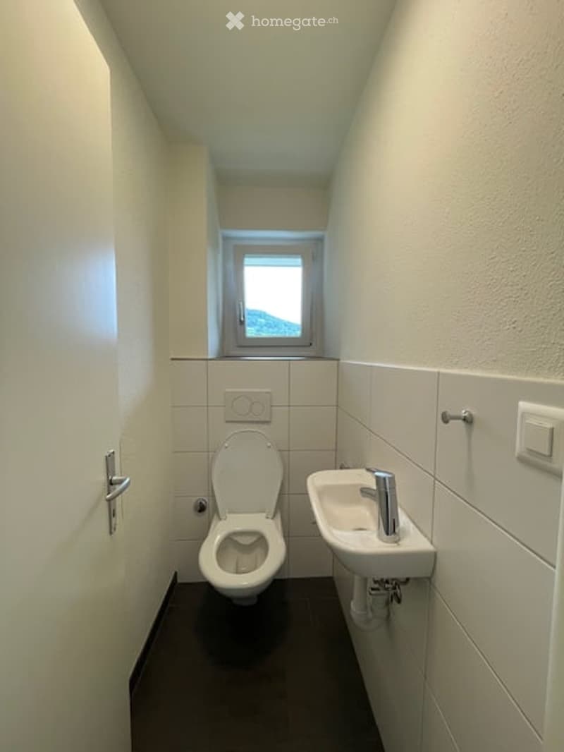 Modern bathroom with white tiled walls, a toilet, a sink with a chrome faucet, and a small window offering a view.
