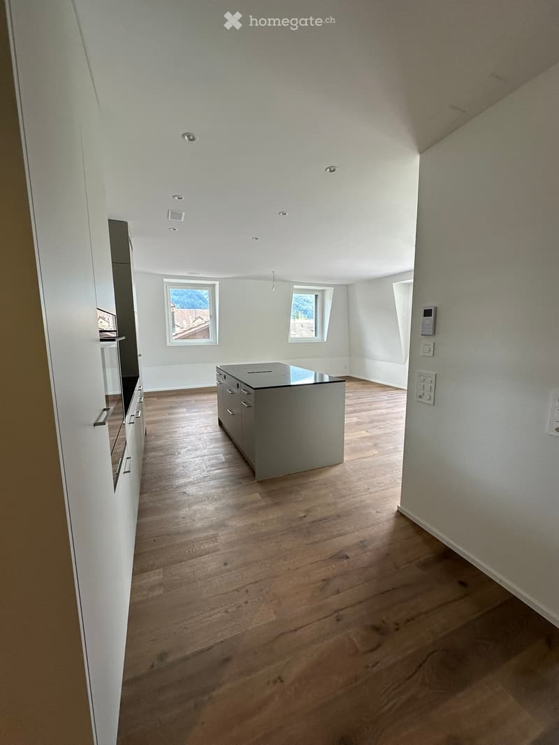 Empty room with wooden flooring, white walls, and recessed ceiling lights. Features a central island with drawers and cabinets, and three windows allowing natural light.