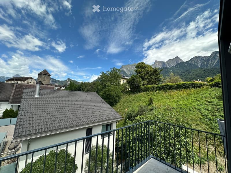 Balcony overlooking a scenic view of a town with houses, trees, and distant mountains under a partly cloudy sky.