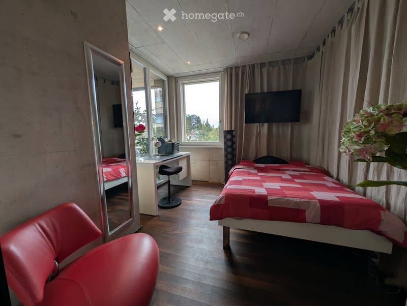 Modern bedroom with two single beds, red leather chair, large mirror, desk with stool, and a TV screen above the window.