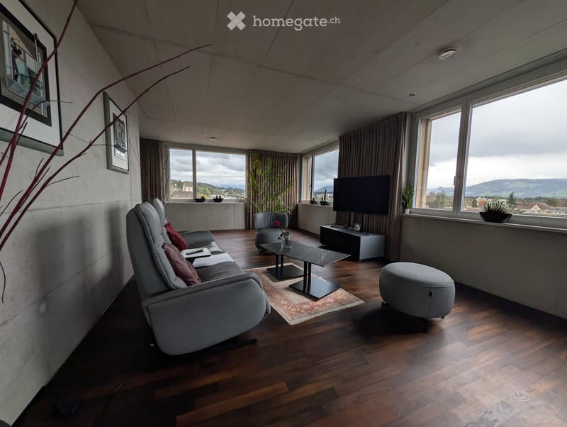 Modern living room with wooden flooring, large windows offering a scenic view, a grey sofa, coffee table, ottoman, armchair, and a television.