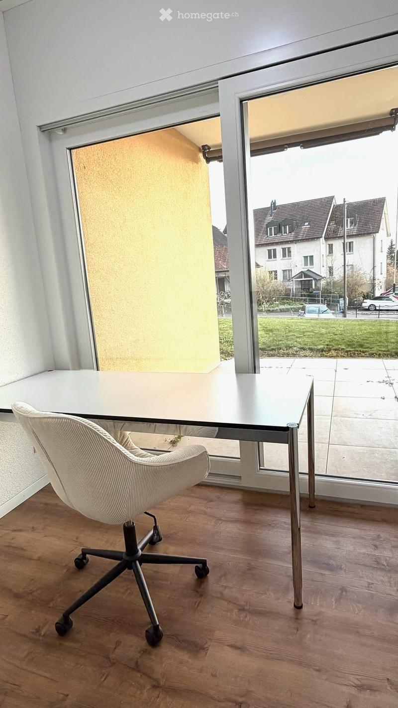 A modern, minimalist room with a white table and a single white chair, large windows offering a view of the outside, and a wooden floor.