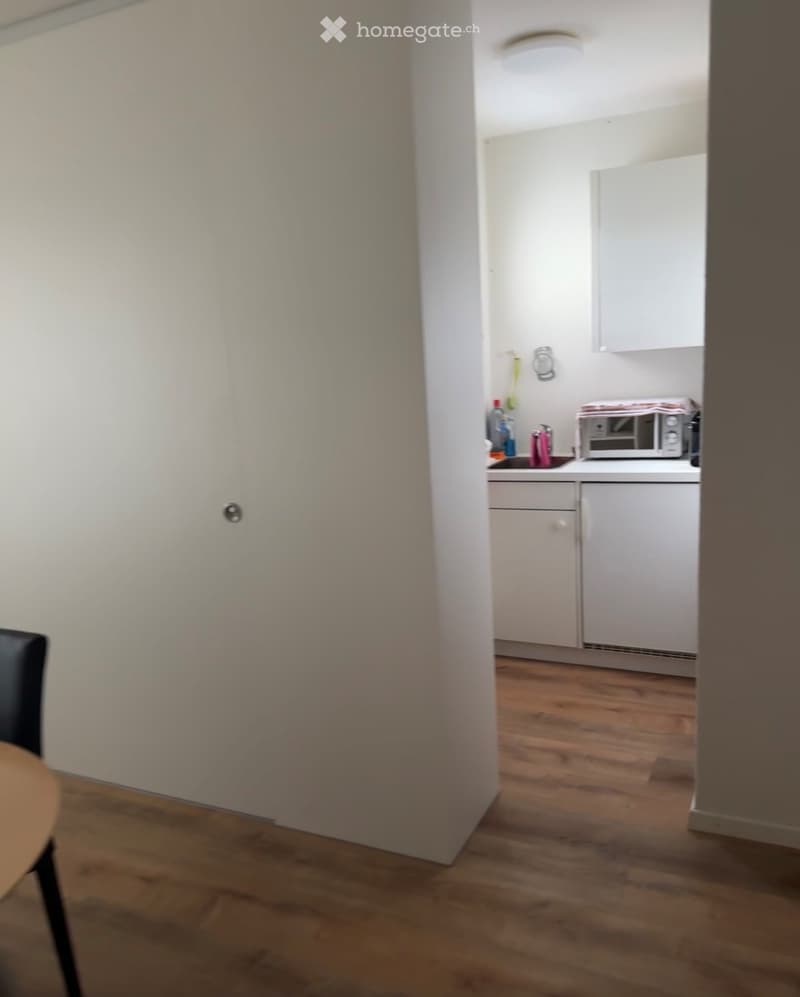 A small, modern kitchen with white cabinets, a microwave, and a sink. The adjacent room features a wooden floor, a chair, and a small table.