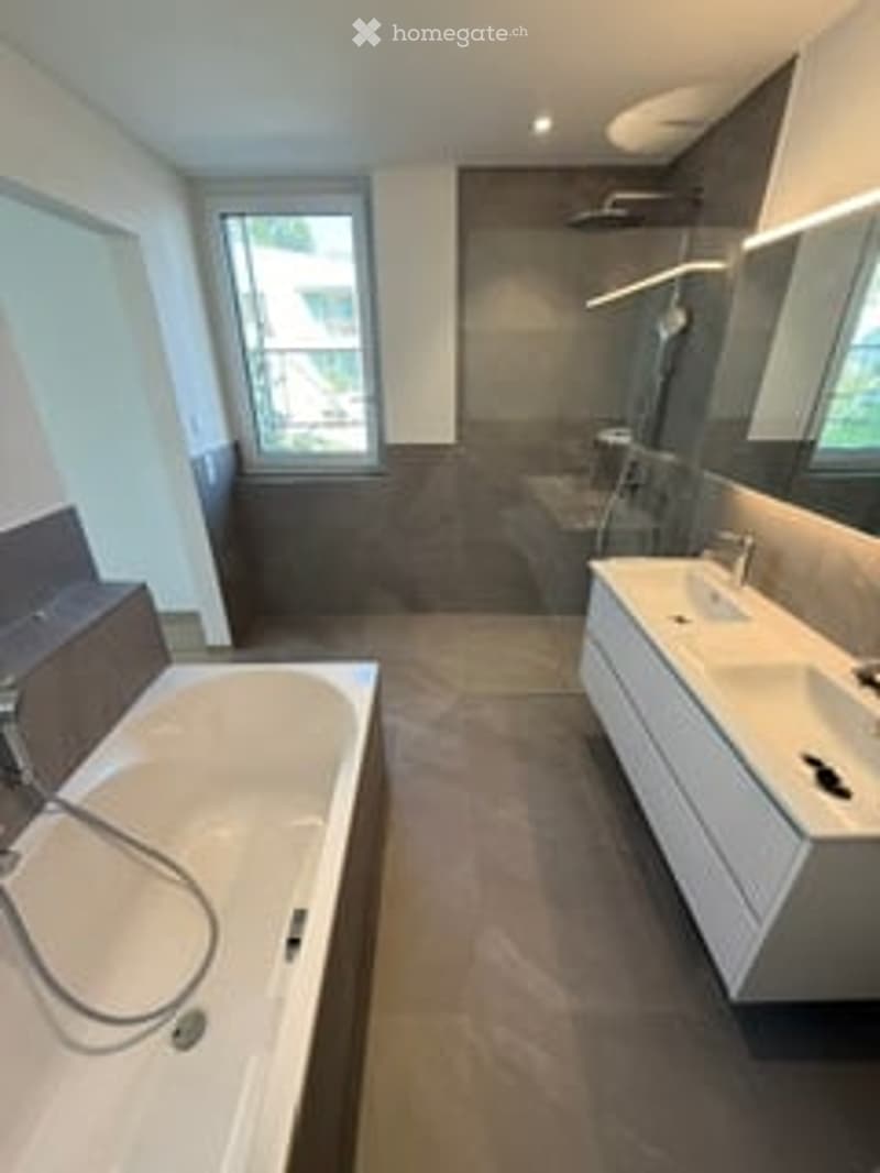Modern bathroom with a large bathtub, double sink, and large mirror. The space features dark tiles and a sleek, contemporary design.