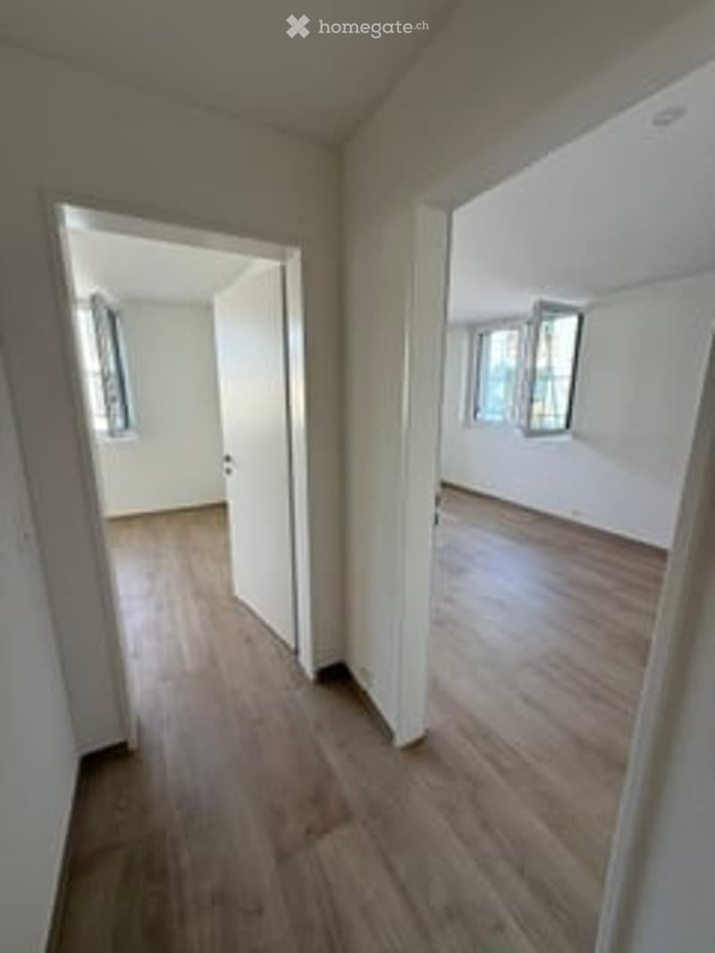 Empty room with wooden flooring and white walls, featuring large windows and an open doorway leading to another room.