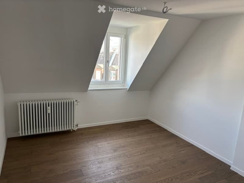 Empty attic room with wooden flooring, white walls, and a single window with a slanted roof design. A radiator is visible against one wall.