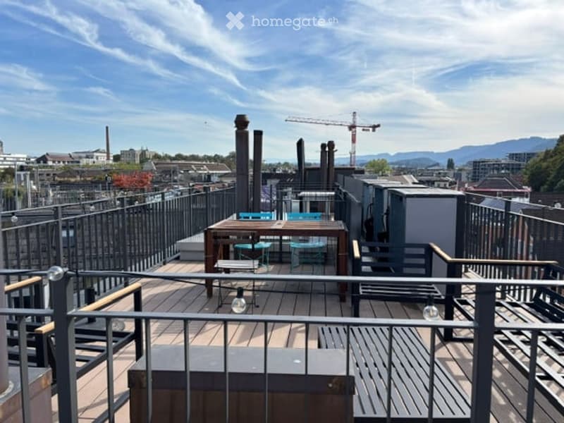 Rooftop terrace with wooden decking, metal railing, and outdoor furniture including a table and chairs, overlooking an urban landscape with construction cranes and distant mountains.