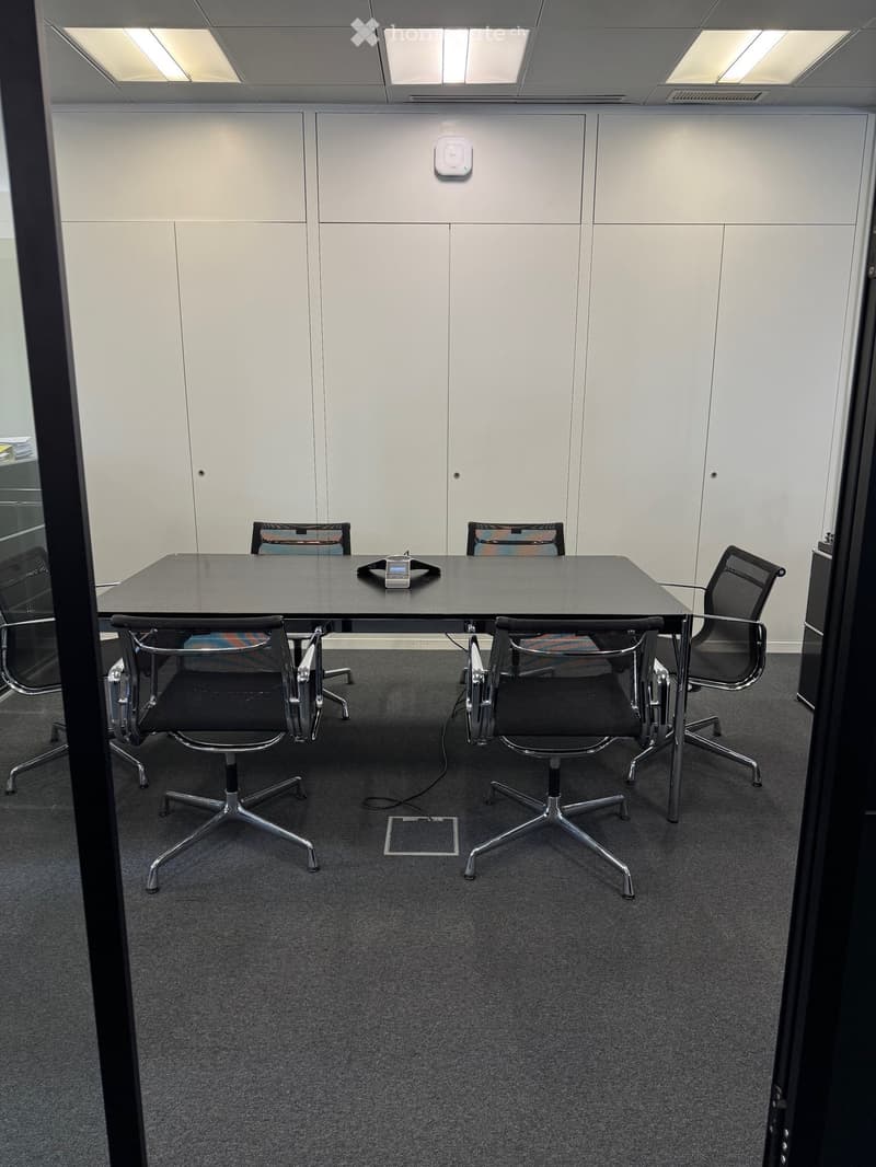 A modern office meeting room with a large black conference table surrounded by ergonomic chairs, white walls, and a carpeted floor.