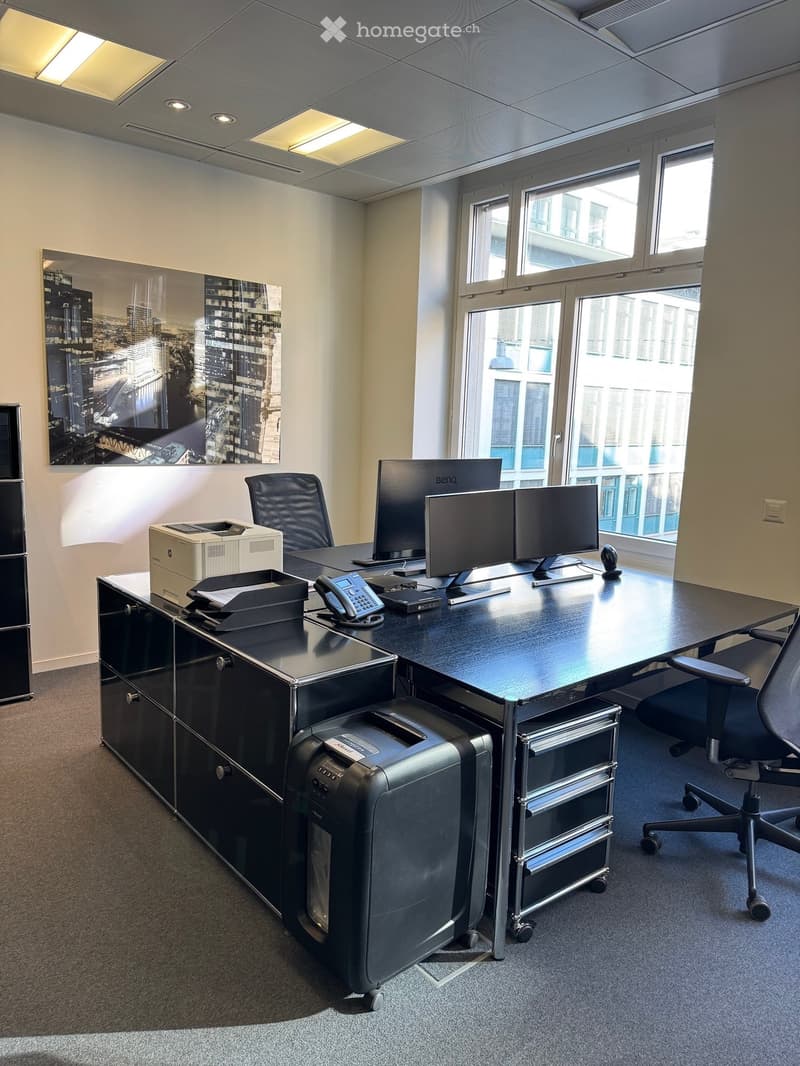 Office space with a large desk, two monitors, a printer, a telephone, and two office chairs. Large windows provide natural light, and a cityscape artwork adorns the wall.