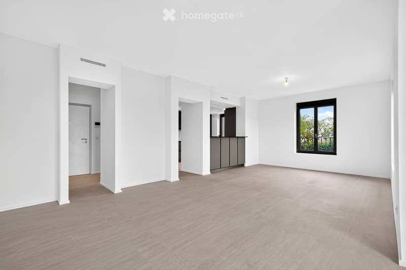 Empty room with white walls, light wooden flooring, and a large window with a view of greenery. The room features a doorway leading to another area and a small kitchen area visible in the background.