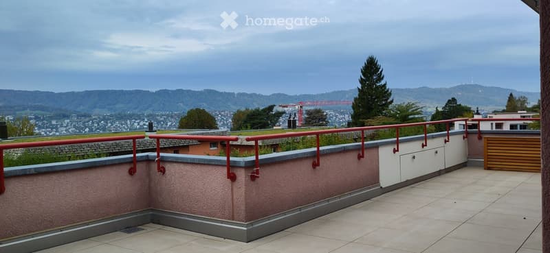 Roof terrace with red metal railing, tiled floor, and scenic city and mountain view.