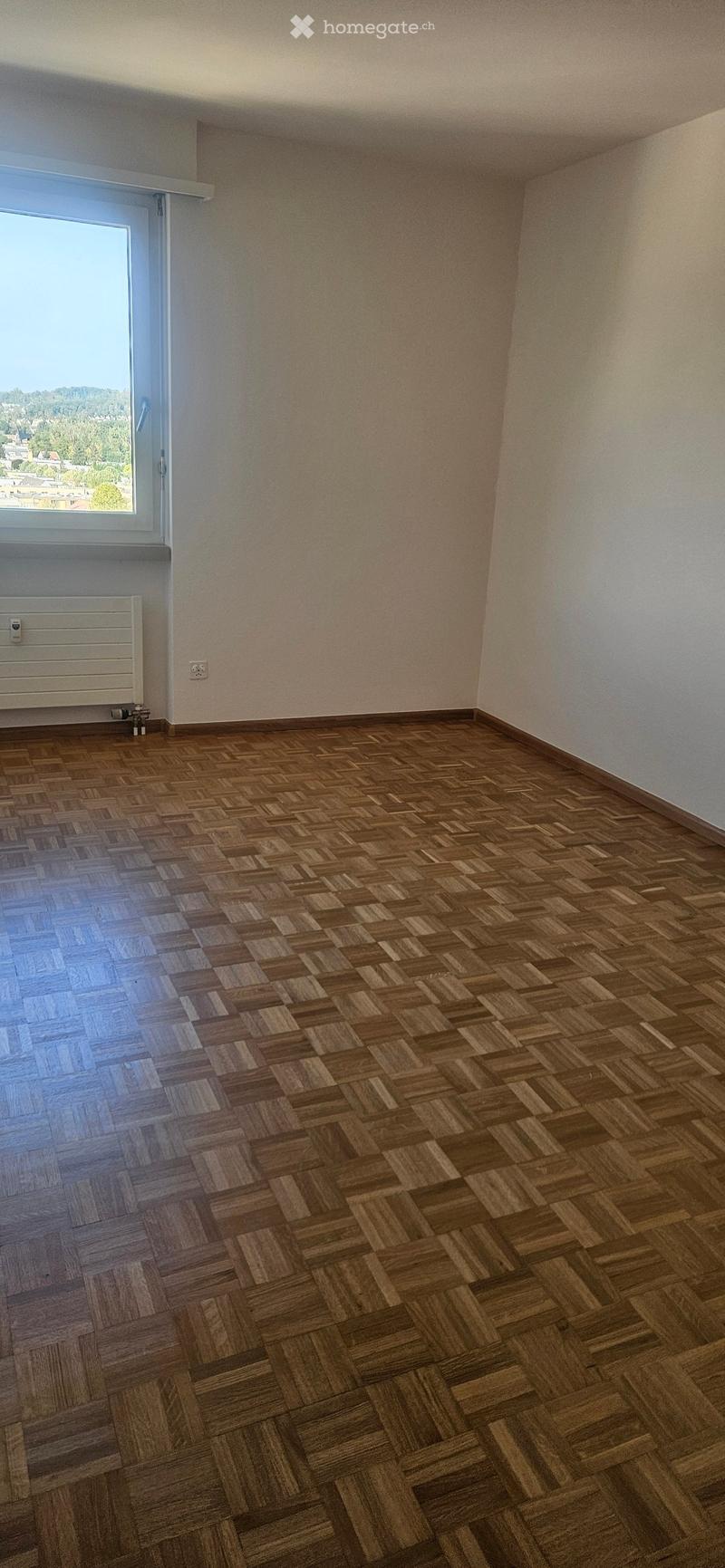 Empty room with wooden herringbone flooring, white walls, and a large window with a view of the landscape.