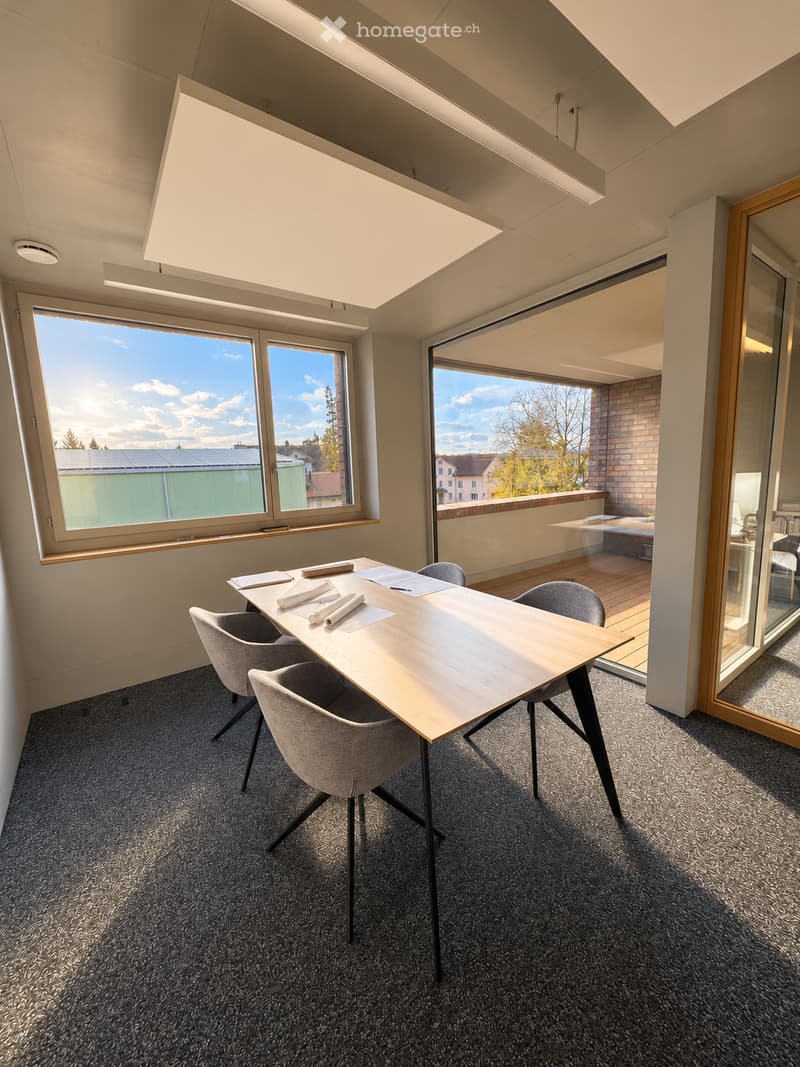 Modern office or meeting room with a large wooden table, four grey chairs, and large windows offering a view of the outside. The room features a grey carpeted floor and a sleek, contemporary design.