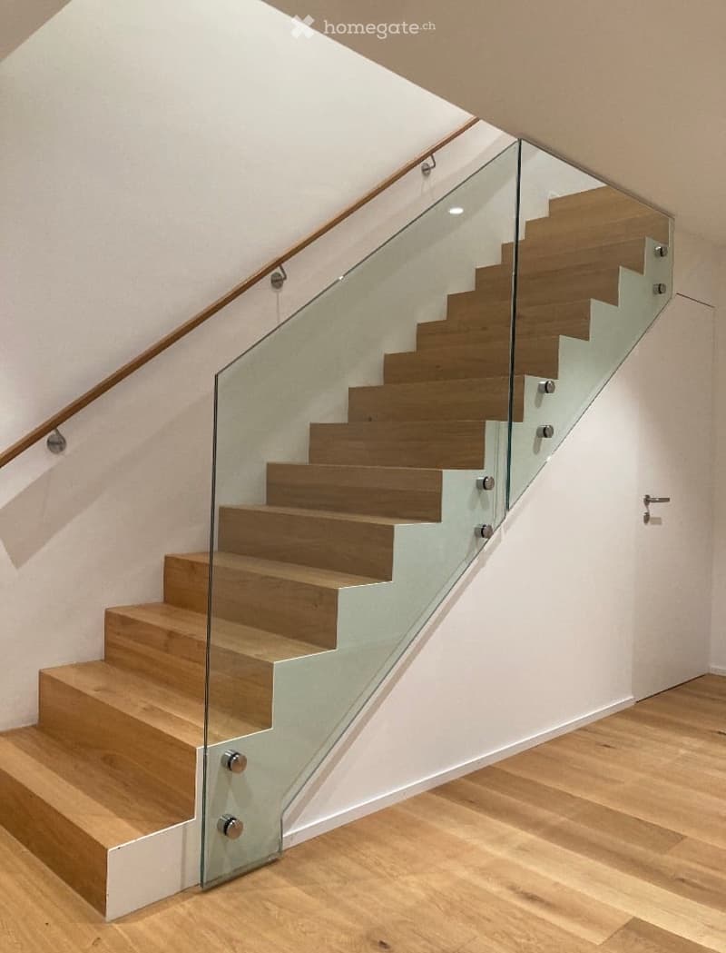 Modern staircase with wooden steps and glass railing, set in a bright hallway with light-colored walls and wooden flooring.