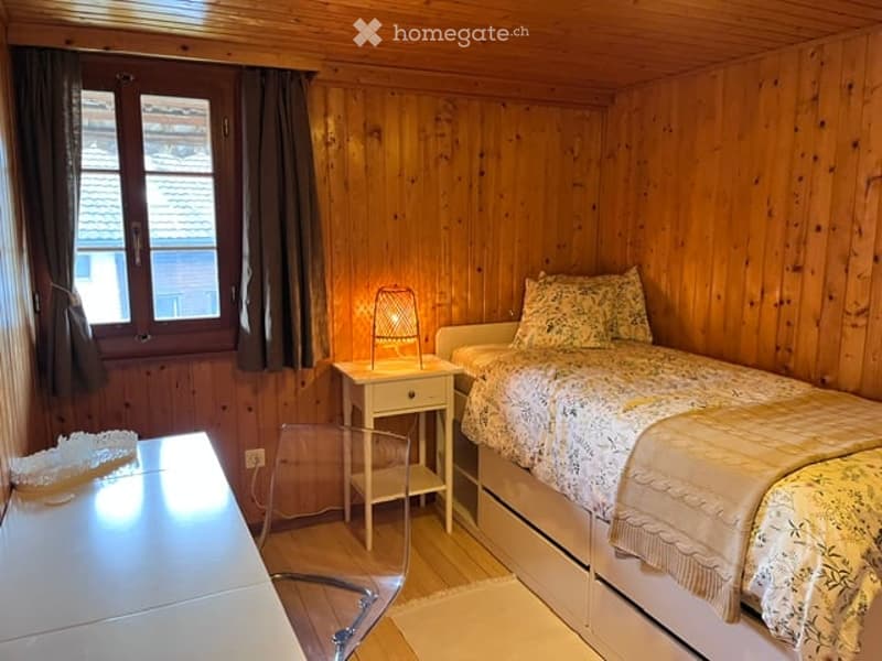 Cozy wooden bedroom with a single bed, nightstand, lamp, and a small chair. The room features wooden walls and a window with curtains.