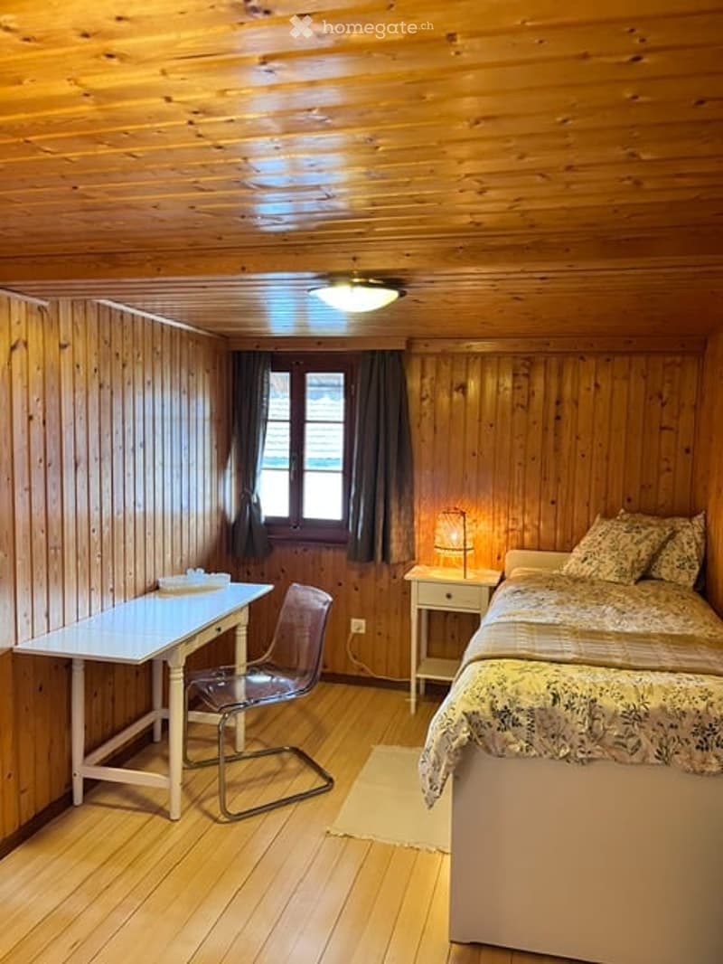 Cozy wooden bedroom with a double bed, nightstand with lamp, small desk, and chair. Wooden walls and ceiling, warm lighting, and a window with dark curtains.