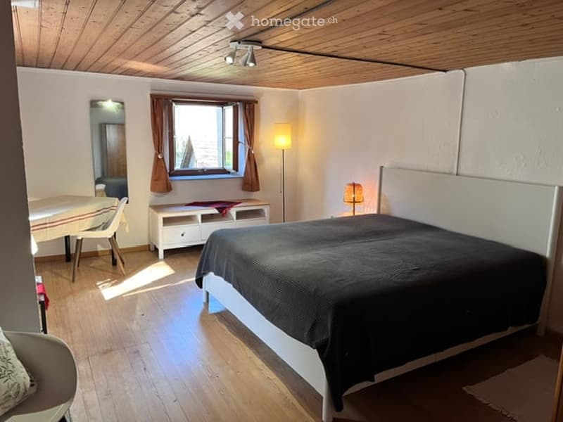 Cozy bedroom with wooden floors, white walls, and a slanted wooden ceiling. Features a large bed with dark bedding, a small table with chairs, a mirror, and a floor lamp near a window with brown curtains.
